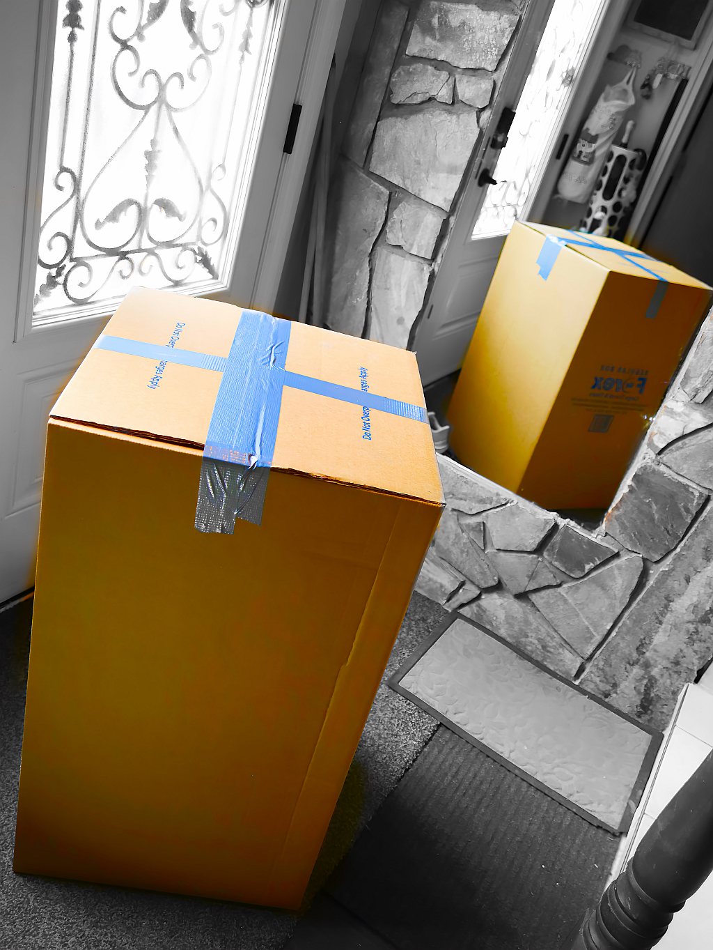A Balikbayan box waiting to be picked up by the front door and standing beside a tall mirror with its reflection on it.  Balikbayan box has a very saturated yellow-orange color while it has a black & white background.